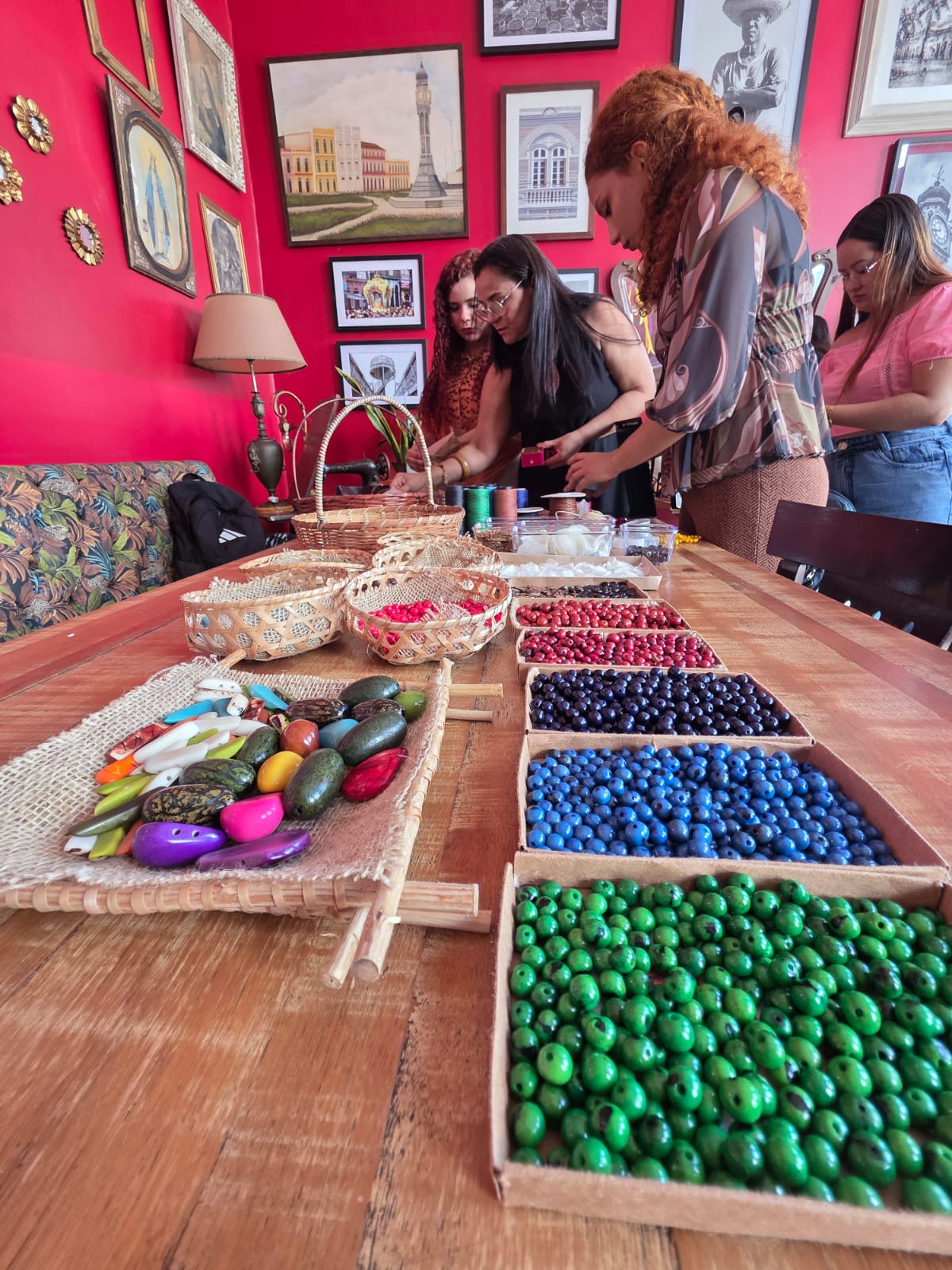 Mulheres manuseiam materiais de artesanato sobre uma mesa comprida de madeira. Em primeiro plano, destacam-se caixas organizadoras enfileiradas com sementes e miçangas coloridas em tons de verde, azul, roxo e vermelho.