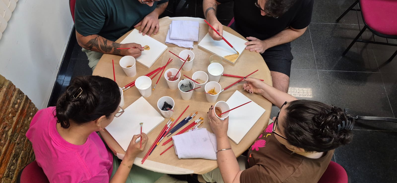 Visão aérea de quatro pessoas sentadas ao redor de uma mesa redonda pintando pequenas telas brancas com pincéis de cabo vermelho. No centro da mesa, há copos descartáveis com tintas em tons terrosos.