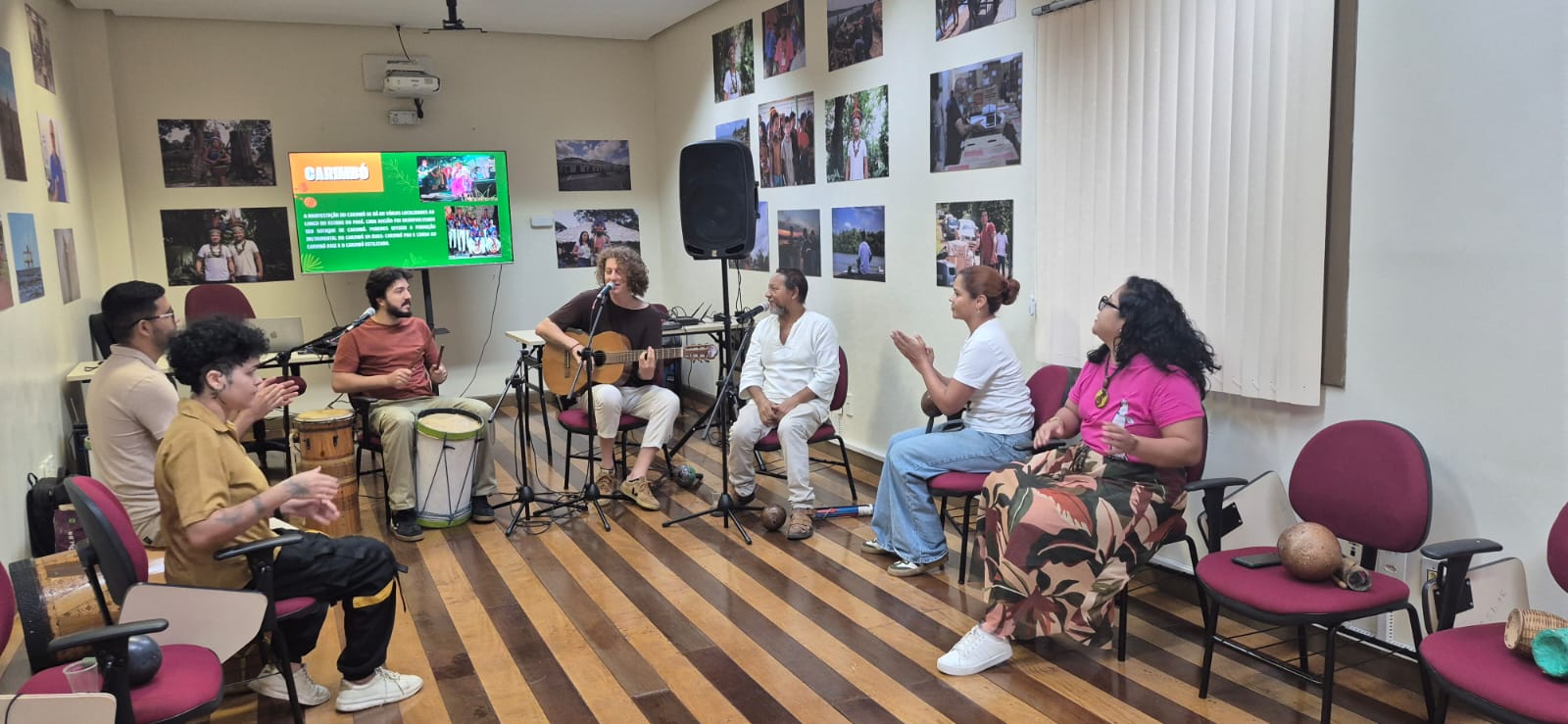 Grupo de pessoas reunido em círculo em uma sala com piso de madeira, tocando instrumentos e cantando. Ao fundo, uma televisão exibe um slide verde com a palavra 'CARIMBÓ' em letras amarelas.