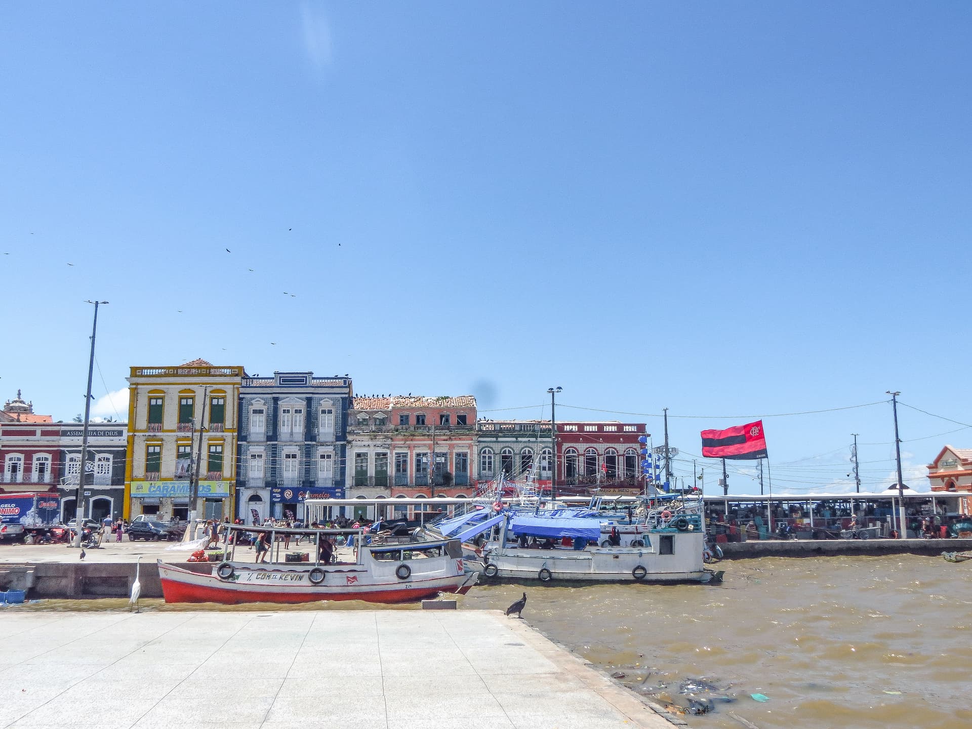 Embarcações de madeira atracadas em um cais de águas turvas. Ao fundo, ergue-se uma fileira de casarões históricos coloridos em tons de amarelo, azul, rosa e vermelho, sob um céu azul e limpo.