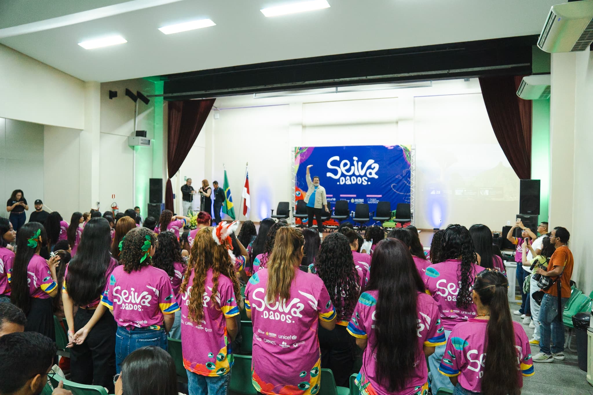 Público de costas, vestindo camisetas rosa com a estampa 'Seiva.dados', assiste a uma apresentação em um auditório. No palco ao fundo, um palestrante ergue o braço diante de um painel azul com o mesmo logotipo do evento.
