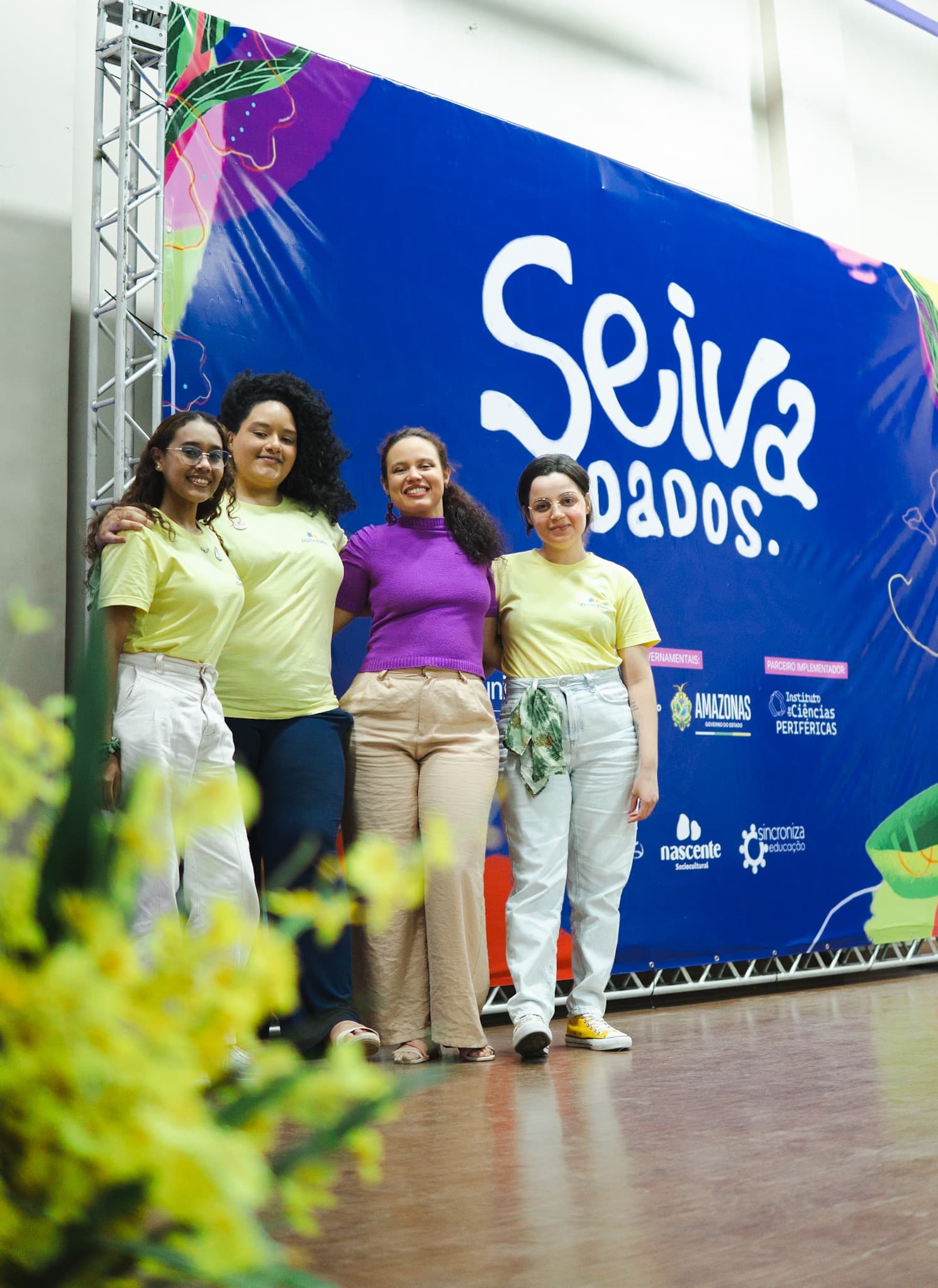 Quatro mulheres sorriem abraçadas em um palco, posicionadas à frente de um grande painel azul com a inscrição 'Seiva.dados'. Três delas vestem camisetas amarelas claras e uma veste blusa roxa.