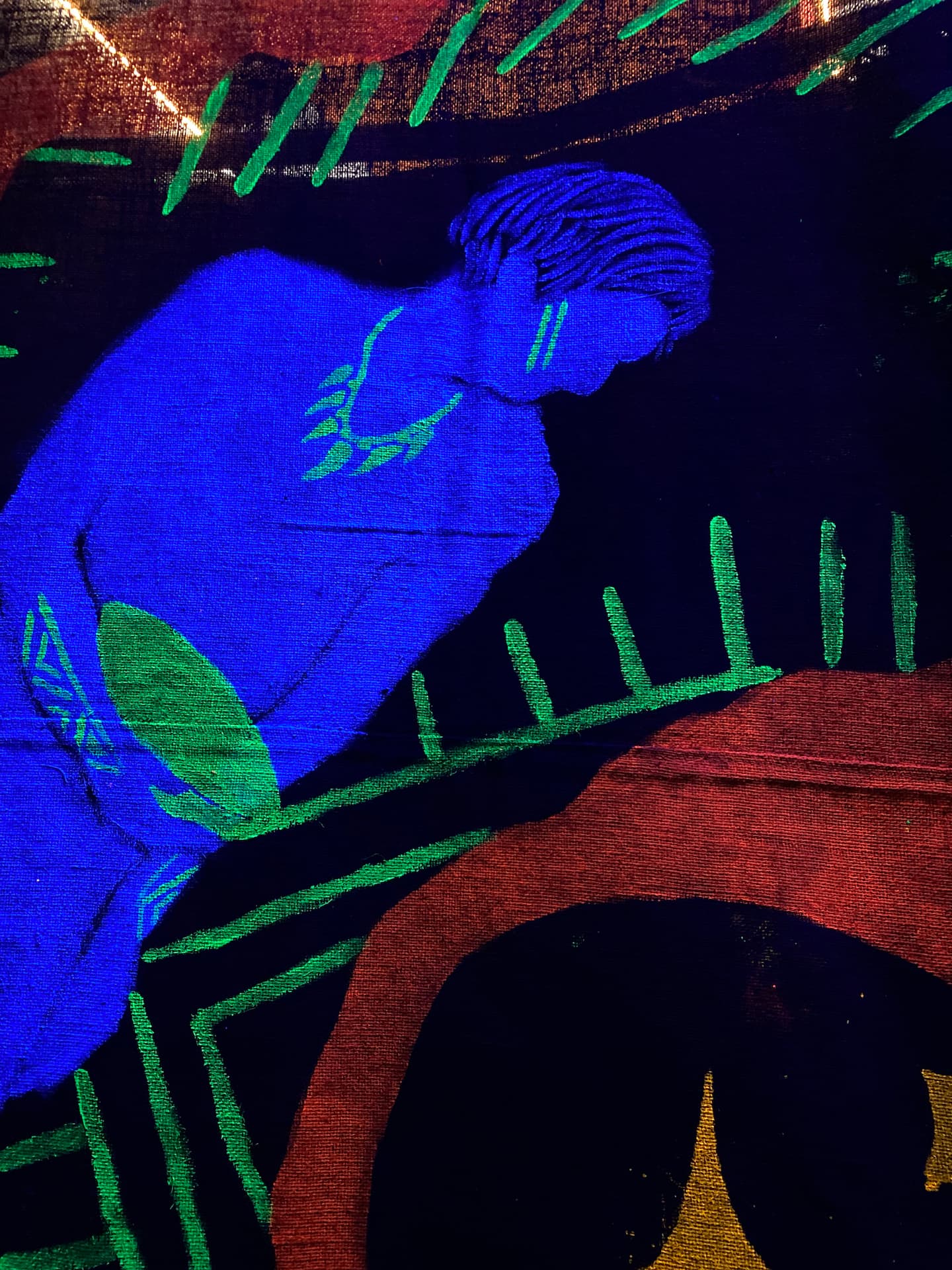 Pintura em tecido de uma figura indígena estilizada em tons de azul brilhante, com traços verdes na bochecha, pescoço e perna. O fundo é escuro, com grafismos em verde, vermelho e amarelo.