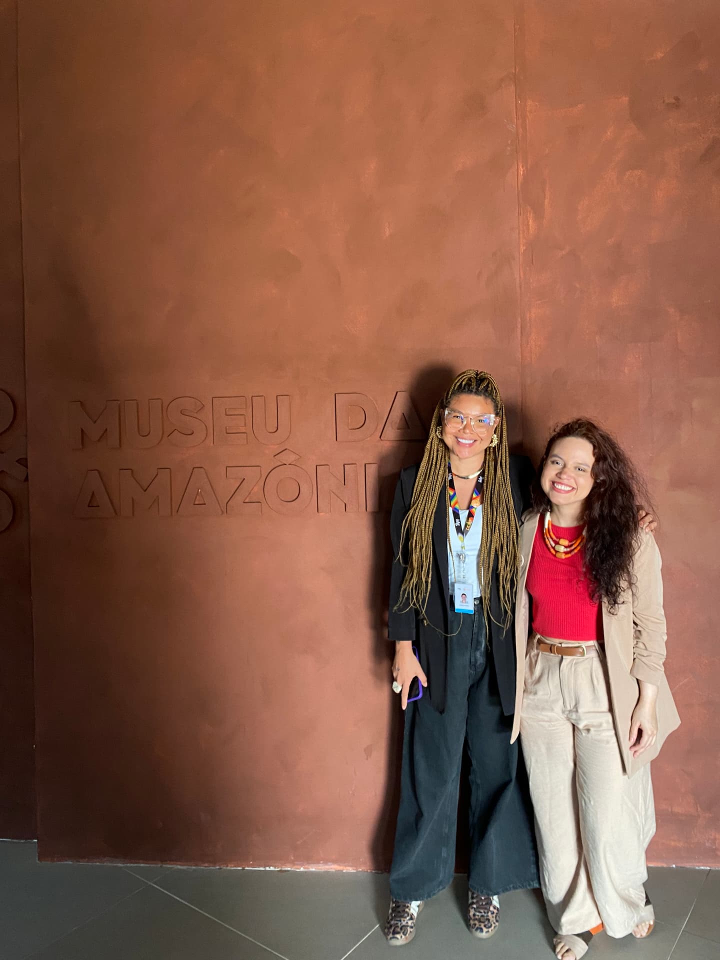 Duas mulheres sorriem abraçadas à frente de uma parede marrom-terrosa com a inscrição em relevo 'MUSEU DA AMAZÔNIA'. A mulher à esquerda usa tranças longas e blazer preto, e a da direita veste blazer bege e blusa vermelha.