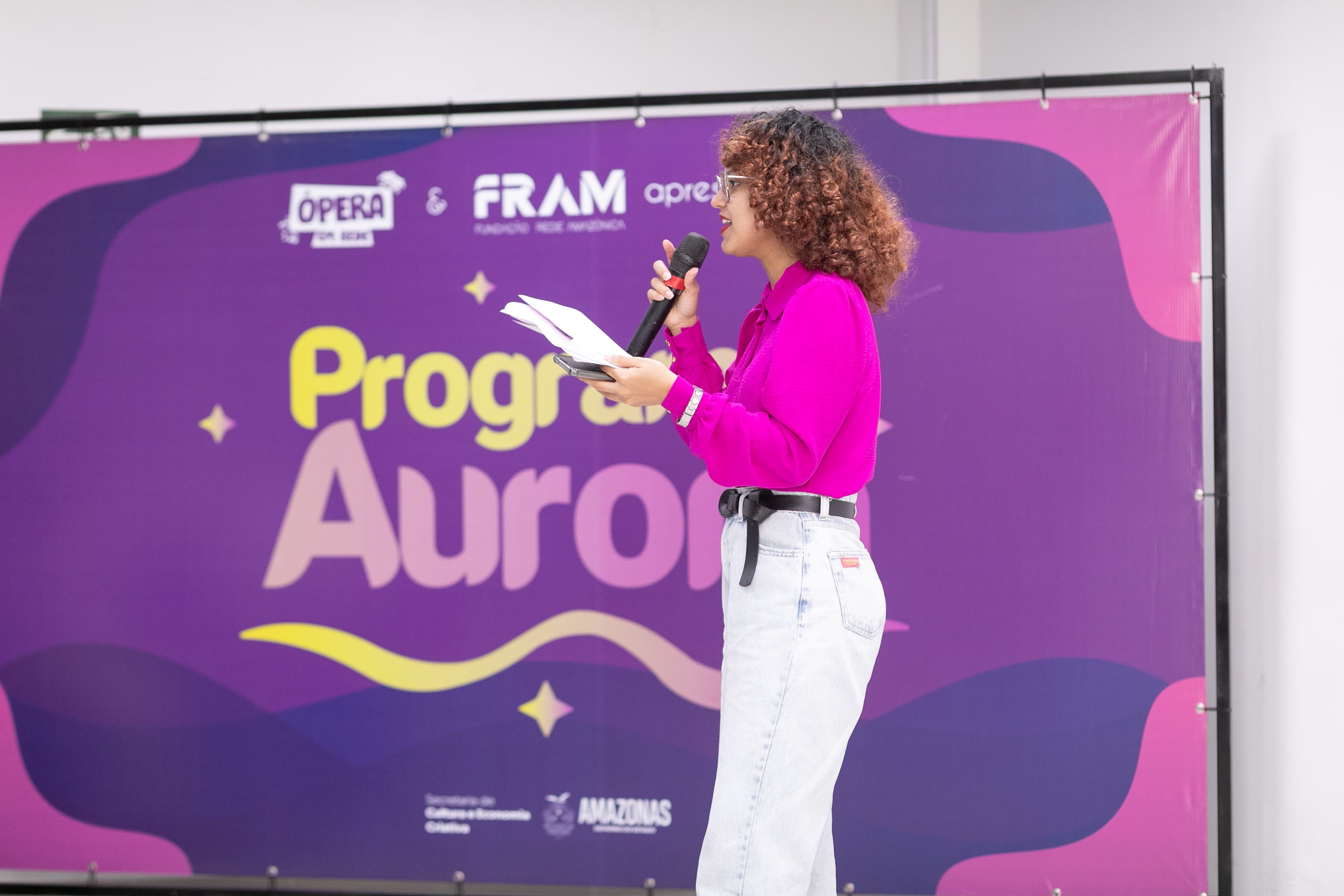 Uma mulher de cabelos cacheados e blusa rosa choque fala ao microfone enquanto segura papéis. Ao fundo, há um painel roxo com a escrita 'Programa Aurora'.