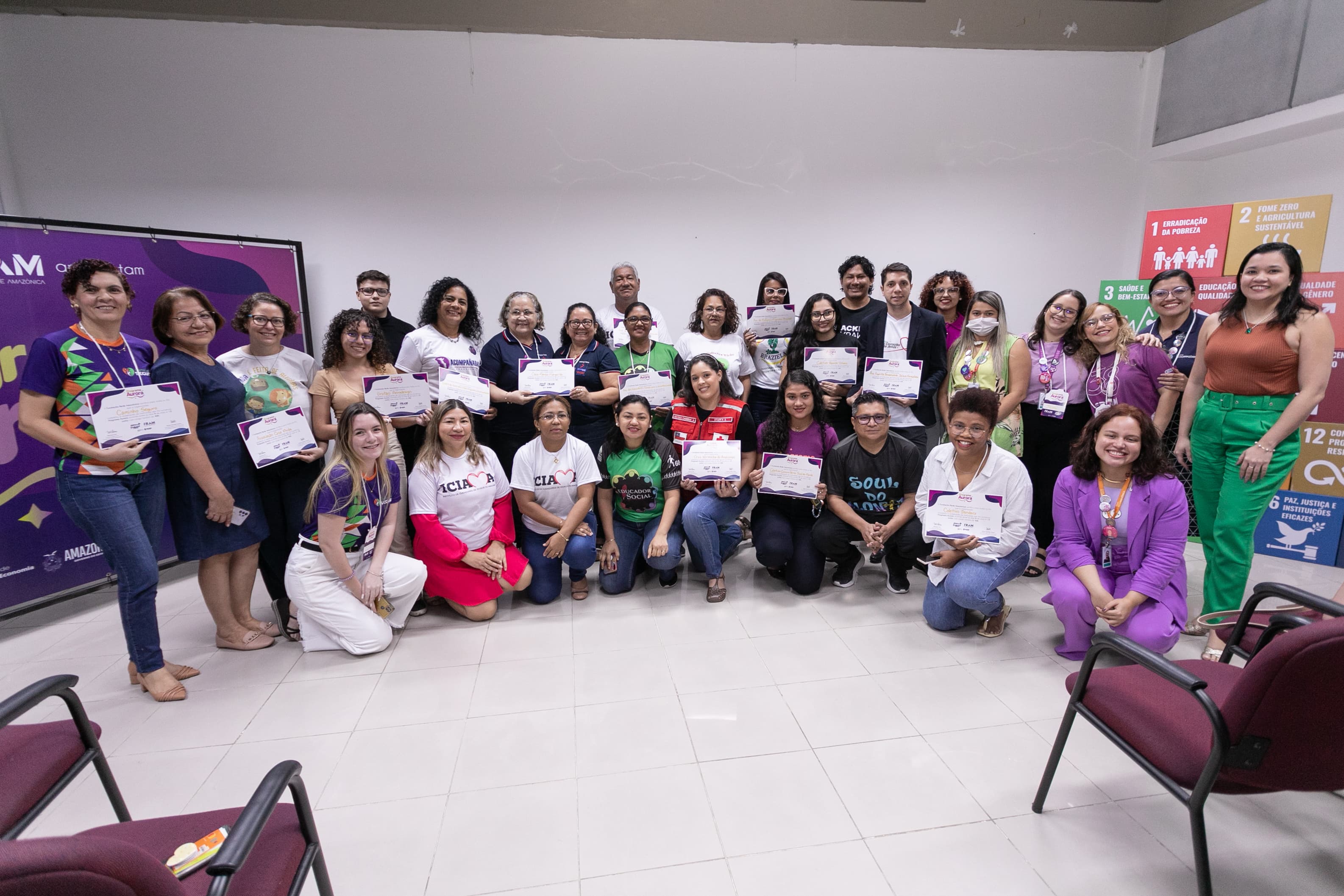 Um grande grupo de pessoas posa reunido em uma sala clara para uma foto coletiva. Muitas delas estão em pé ou agachadas segurando certificados, e a parede ao fundo exibe cartazes coloridos dos Objetivos de Desenvolvimento Sustentável (ODS).