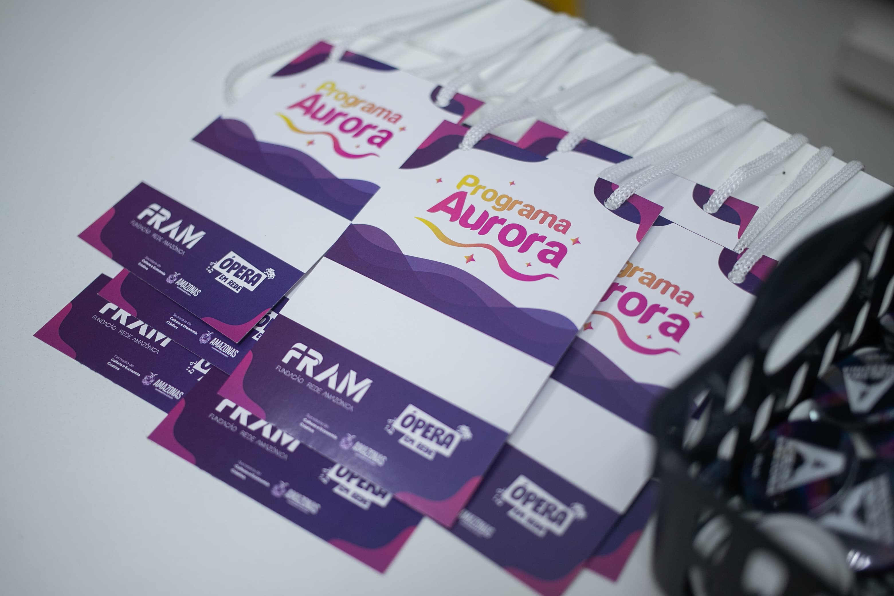 Crachás de identificação brancos e roxos com a inscrição 'Programa Aurora' estão organizados em fileiras sobre uma mesa clara. Ao lado esquerdo, encontram-se pequenos cartões roxos com logotipos do evento.