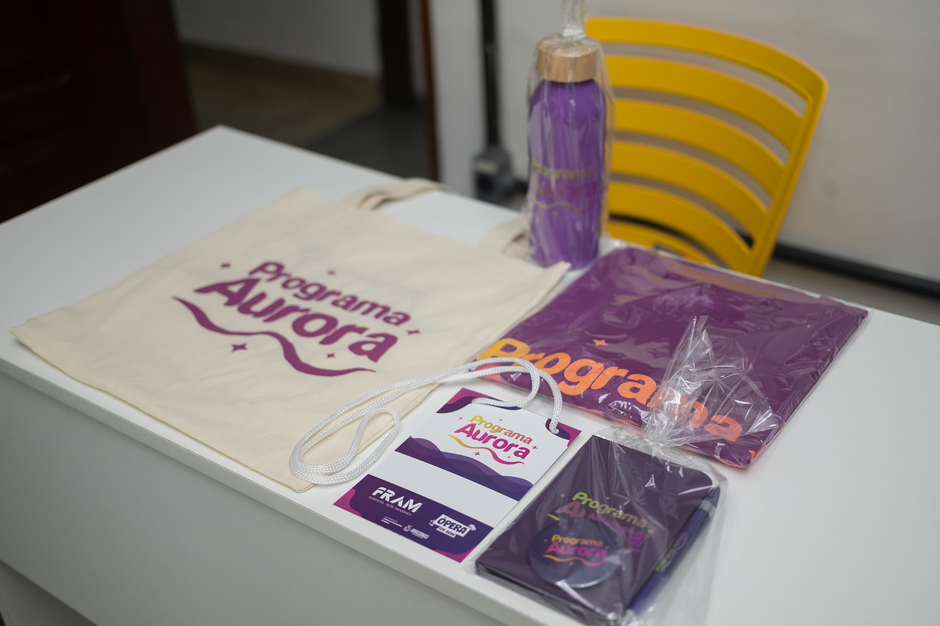 Kit de brindes do 'Programa Aurora' organizado sobre uma mesa branca. Inclui uma sacola ecológica de pano, uma camiseta roxa embalada, uma garrafa térmica roxa, um crachá de identificação e um pequeno bloco de notas.