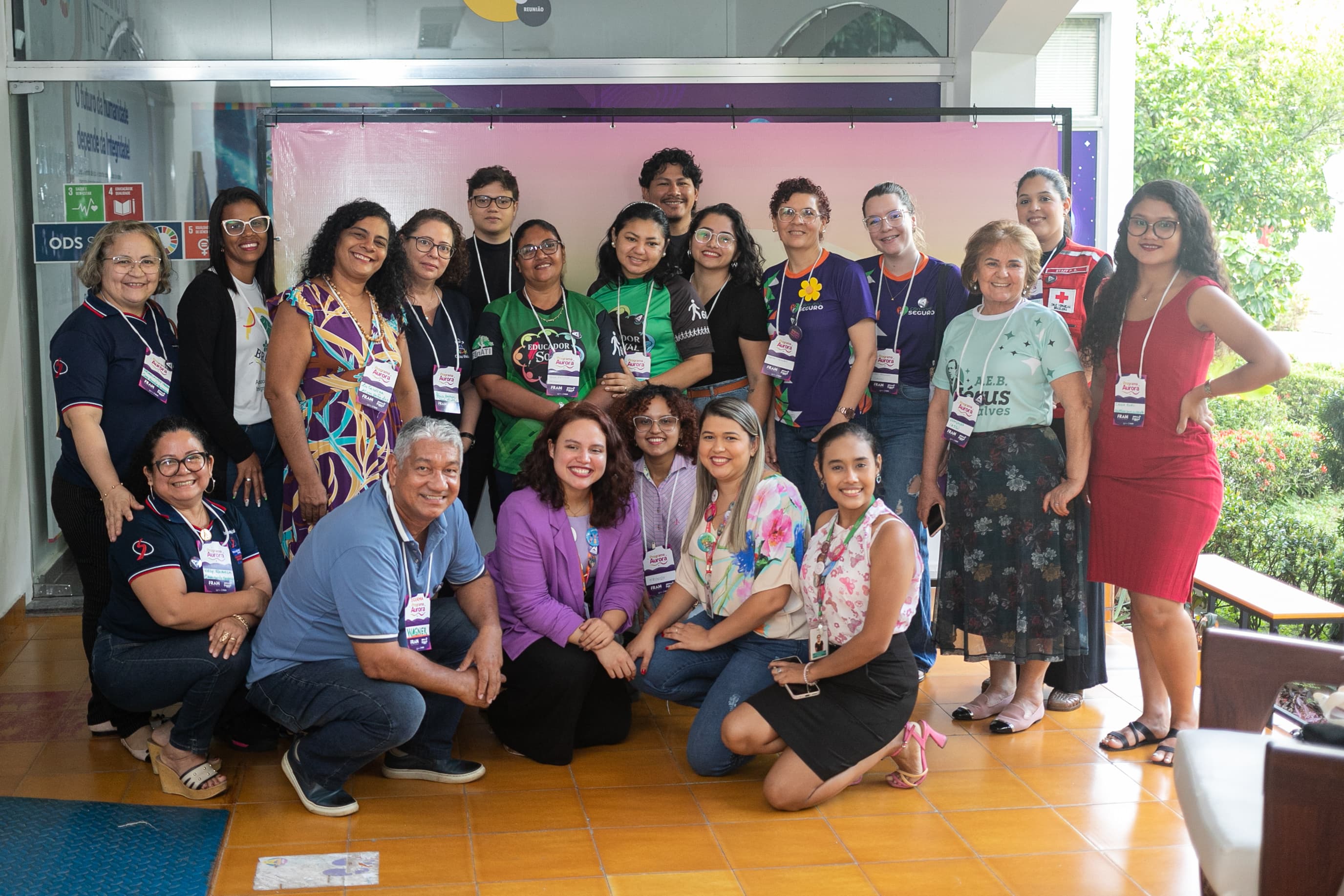 Um grupo de pessoas sorri reunido para uma foto coletiva em uma área coberta com piso cerâmico laranja. Elas usam crachás do evento e estão posicionadas à frente de um painel de fundo em tom rosa-claro.