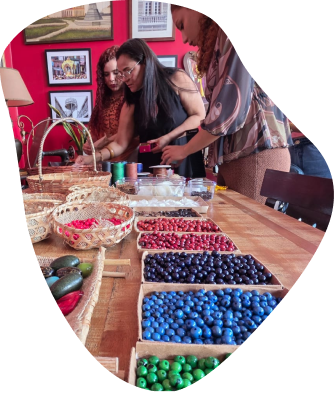 Três mulheres ao redor de uma mesa de madeira repleta de materiais de artesanato. Em primeiro plano, bandejas enfileiradas contêm contas coloridas em vermelho, preto, azul e verde.