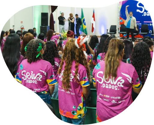 Jovens de costas em um auditório vestindo camisetas rosas com o logotipo 'Seiva.dados'. Algumas usam adornos de penas na cabeça. Ao fundo, pessoas em um palco com bandeiras.